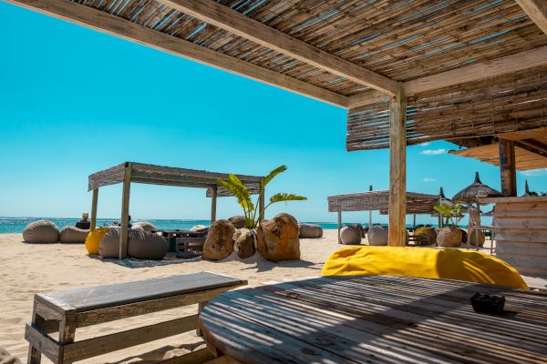 A sunny beach scene featuring shaded wooden structures, benches, and bean bag chairs, with a clear blue sky and ocean in the background.