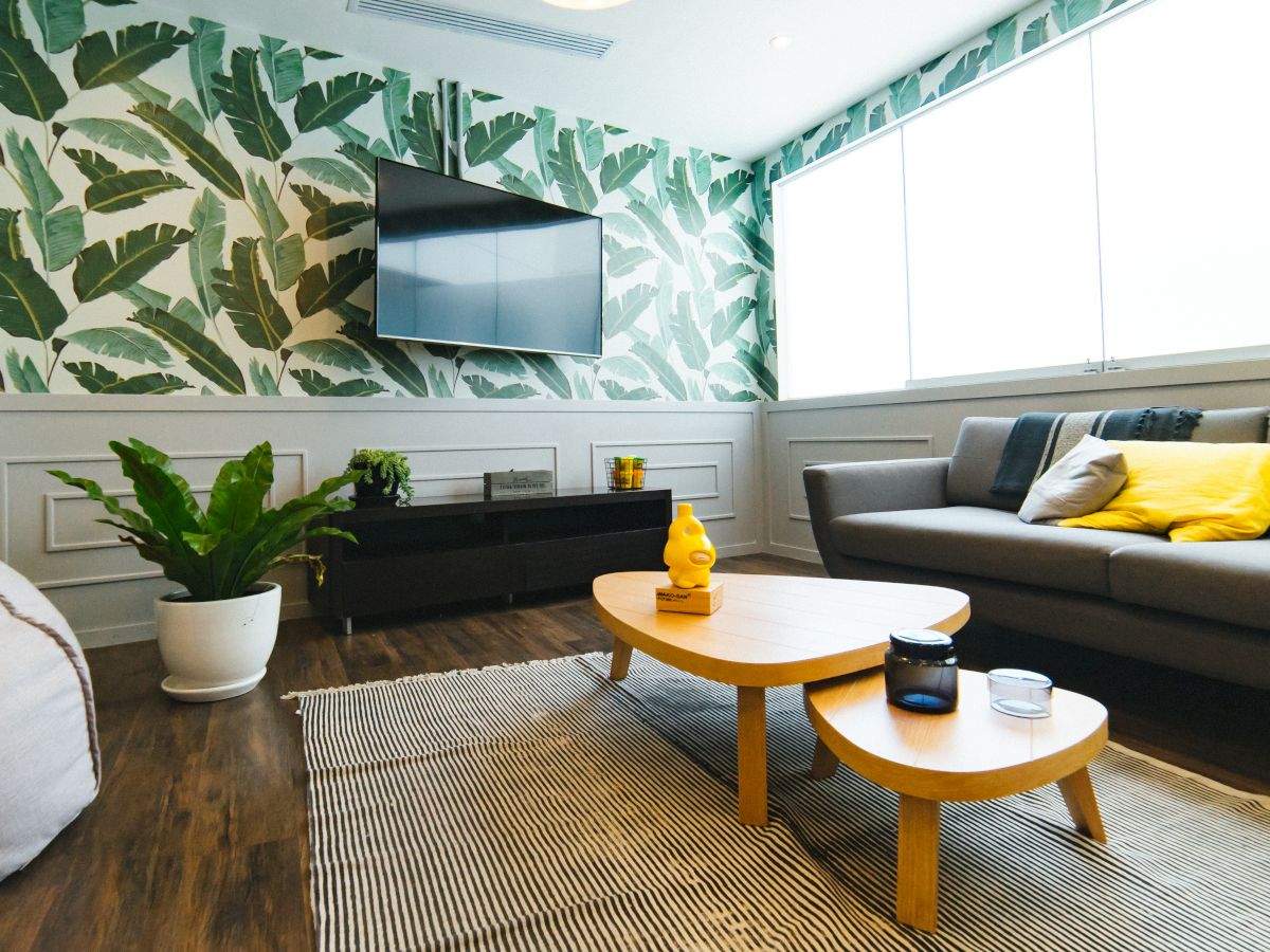 A modern living room with a leafy wallpaper, gray couch, yellow pillows, wooden coffee tables, a rug, TV, and a potted plant.