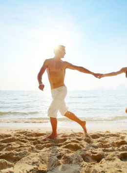 A couple holding hands runs along a sandy beach with the ocean and a bright sun in the background.