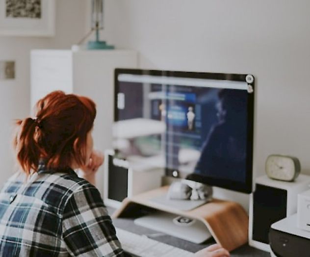 A person with red hair in a plaid shirt sits at a desk working on a computer. The desk has various items including speakers and a clock.
