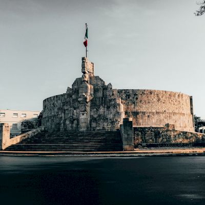 A round stone monument with detailed carvings, prominently featuring a flag on top, stands at the center of a spacious, paved area.