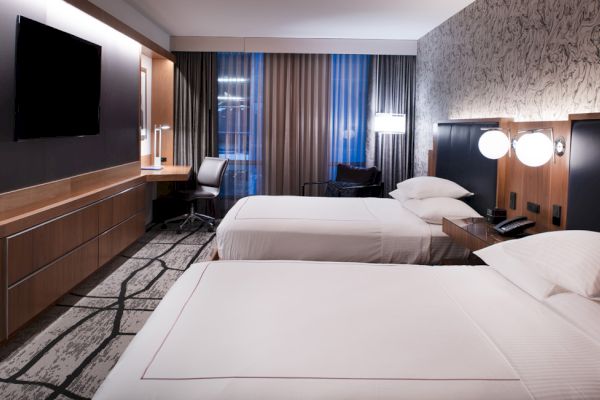 A modern hotel room with two beds, a large TV, a desk, and decorative lighting. The room has a contemporary design with neutral tones.