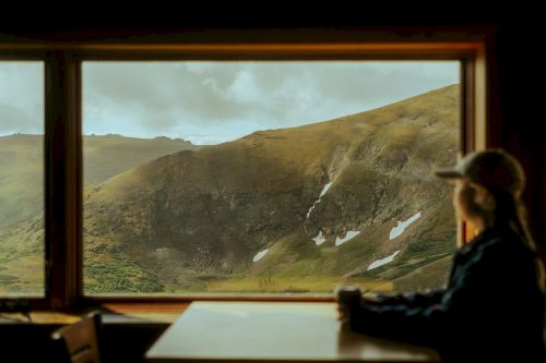 A person is sitting at a table, looking out a large window at a scenic, mountainous landscape with patches of snow.
