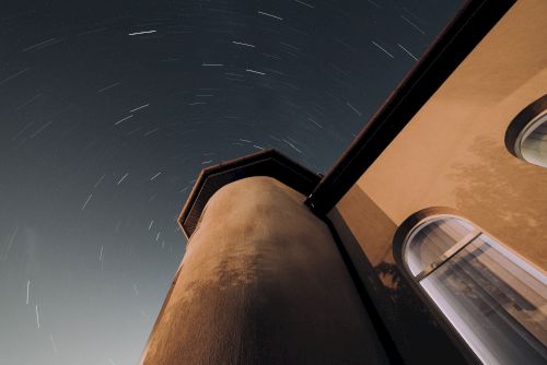 The image shows a building tower at night with a long exposure of stars, creating star trails in the sky. The building has arched windows.