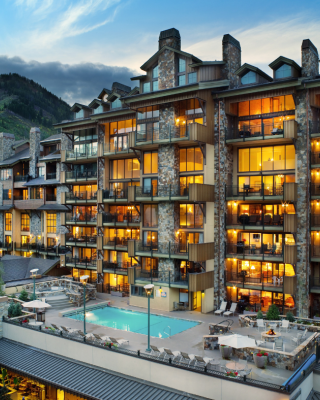 The image depicts a modern hotel with balconies, warm lights, and a poolside area, set against a mountain backdrop at dusk.