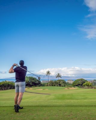 A golfer swings on a lush green course with palm trees and buildings in the background under a bright blue sky.