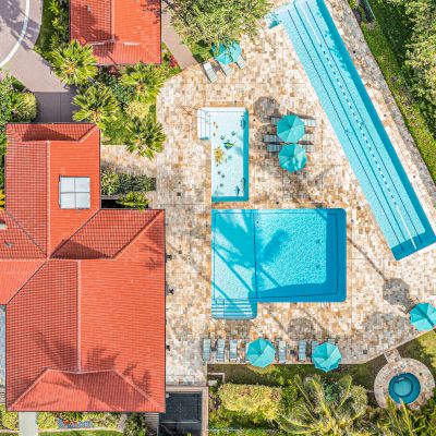 Aerial view of a property with a pool, small outdoor seating areas under umbrellas, and a red-roofed building surrounded by greenery ending the sentence.