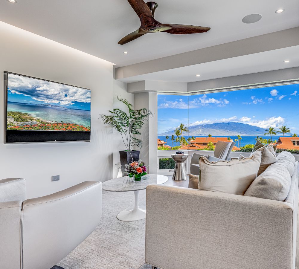 A modern living room with a TV, white furniture, a ceiling fan, and a large window showcasing an ocean view and a clear blue sky dotted with clouds.