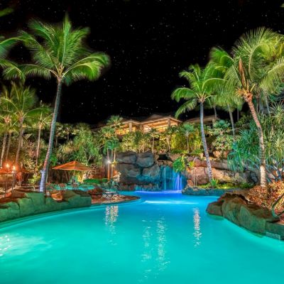 A nighttime scene of a tropical pool lit with blue lights, surrounded by palm trees and rocky landscaping under a starry sky.