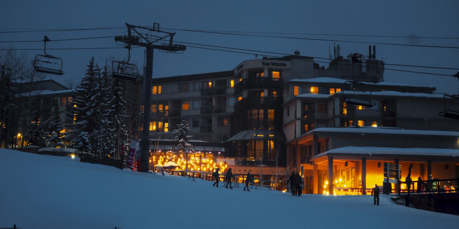 A snow-covered ski resort with illuminated buildings and people skiing at dusk. Ski lift and snowy trees are visible in the background, ending the sentence.