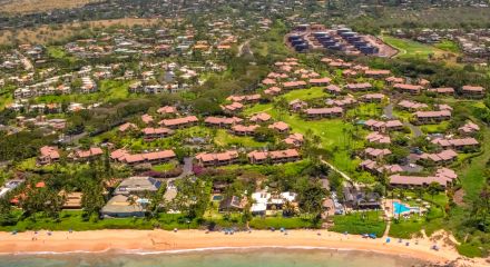 Aerial view of a coastal community with green spaces, residential buildings, and a beach with teal-colored water and some waves along the shore.