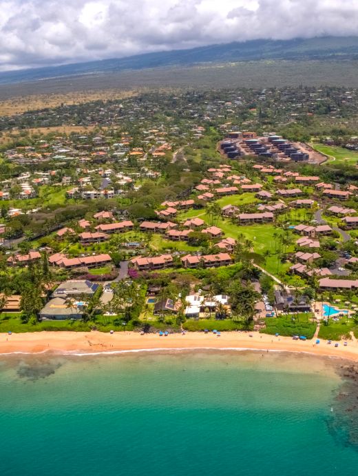 Aerial view of a coastal community with green spaces, residential buildings, and a beach with teal-colored water and some waves along the shore.