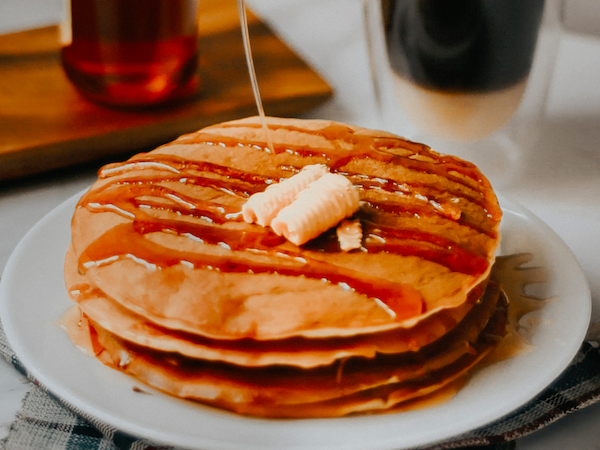 A stack of pancakes with butter and syrup being poured on top, accompanied by a beverage in the background.