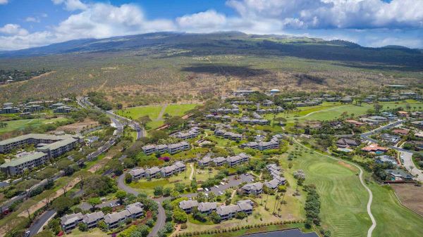 Aerial view of a suburban neighborhood with winding streets, green lawns, a small lake/pond, and surrounding hills under a blue sky.