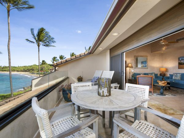 A beachfront patio with white furniture overlooks the ocean, palm trees sway, and an adjacent cozy indoor living space adorned with blue accents.