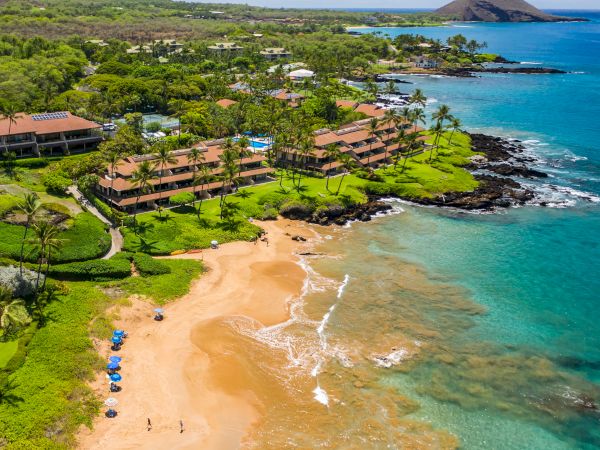 This image shows a tropical beach with clear waters, lush greenery, resort buildings, and a few people and umbrellas on the sand.