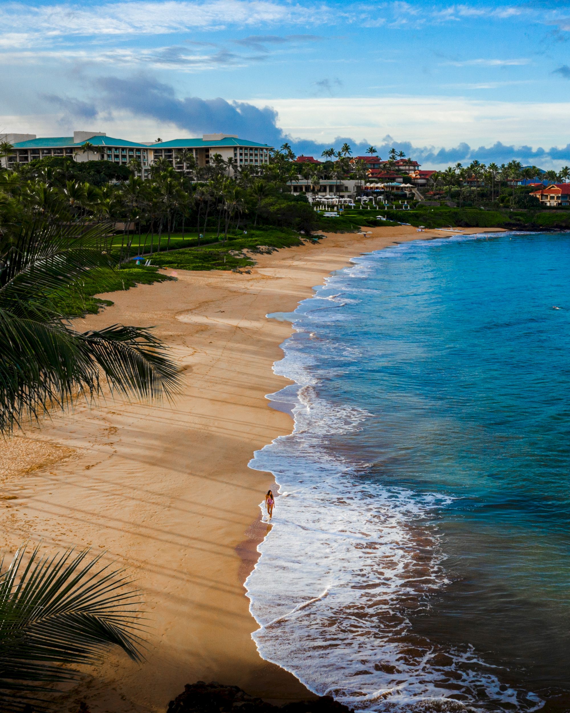 A beach with golden sand and blue water, lined with palm trees and buildings in the background, under a partly cloudy sky.