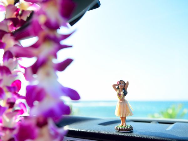 A dashboard hula dancer figurine on a car's dashboard with a blurred lei in the foreground, and a scenic ocean view in the background ending the sentence.