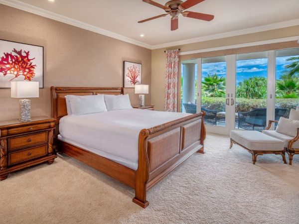 A cozy bedroom with a wooden bed, two nightstands with lamps, artwork, a ceiling fan, and glass doors leading to an outdoor area with lush greenery.