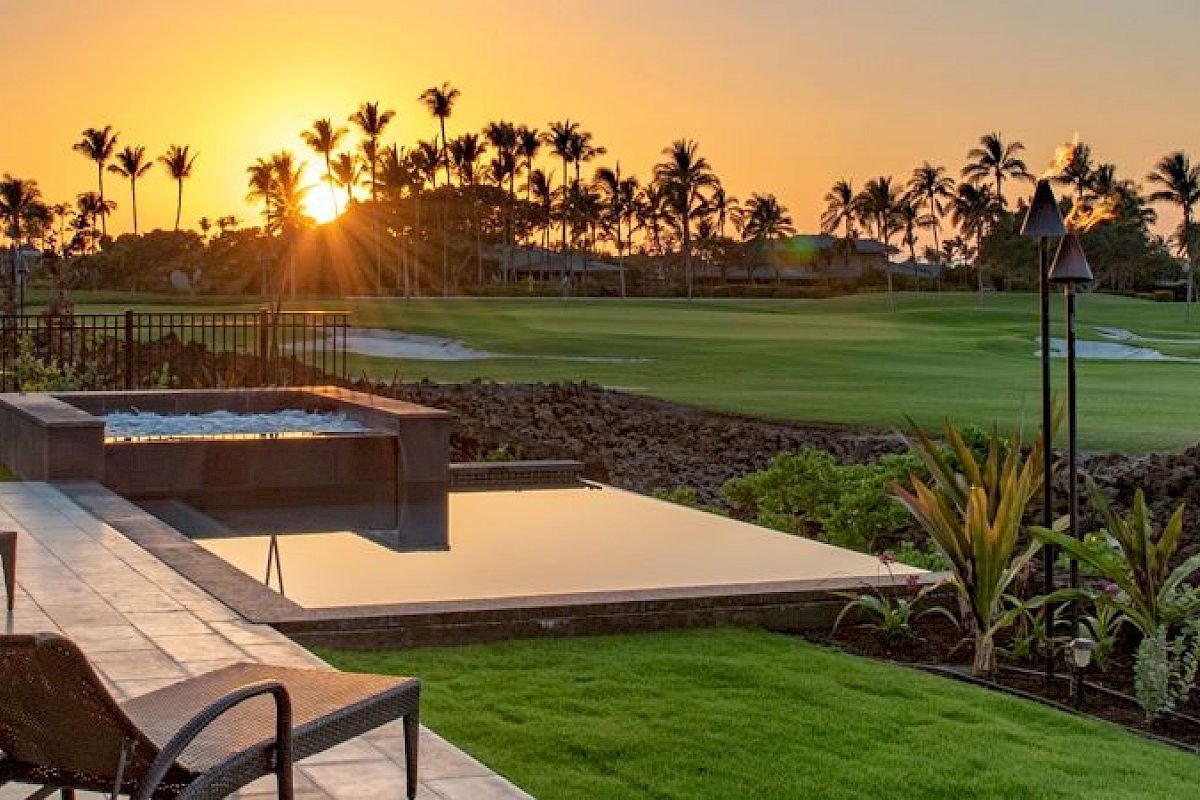 A luxurious patio with lounge chairs, a pool, and a view of a manicured golf course and sunset, surrounded by tropical landscaping and palm trees.