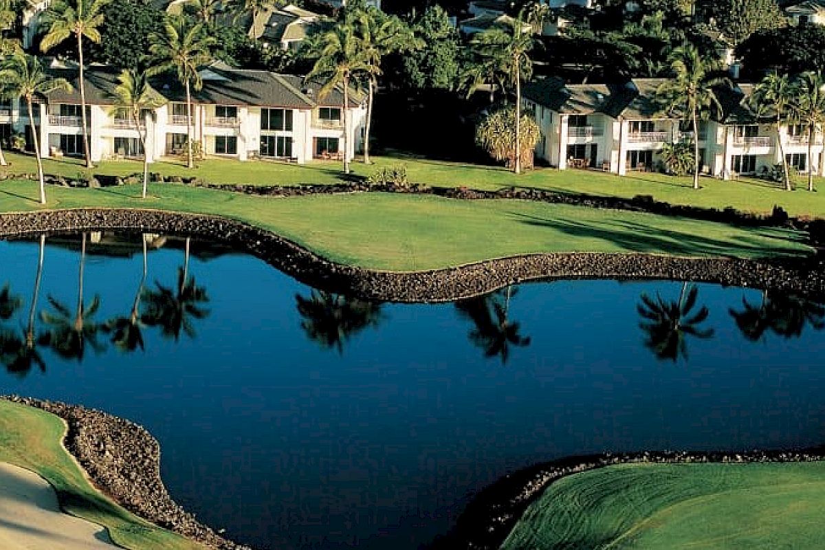 The image shows a picturesque golf course with lush green grass, a heart-shaped water feature, and adjacent residential buildings surrounded by palm trees.