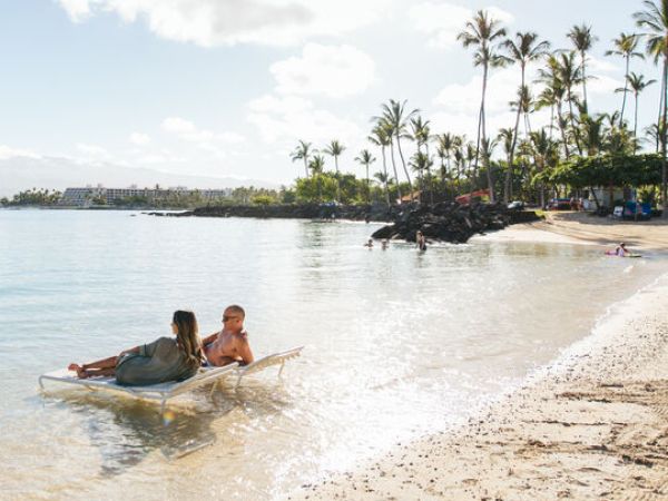 A woman and a child relax on a chair in shallow water at a sunny beach, with palm trees and people in the background.