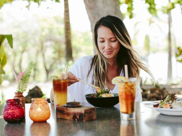 A woman is sitting at an outdoor table, eating food from a bowl, with drinks and decorative items nearby. Trees and greenery are in the background.