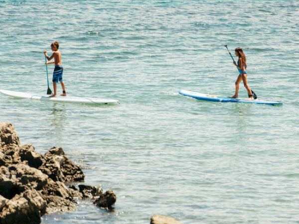 Two people are paddleboarding on the sea near a rocky shore.