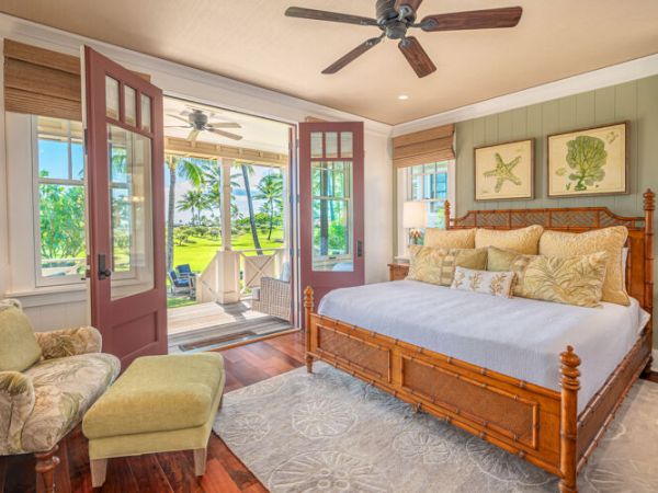 A cozy bedroom with a large bed, armchair, footrest, and two lamps, opens to an outdoor space with chairs and greenery visible through glass doors.