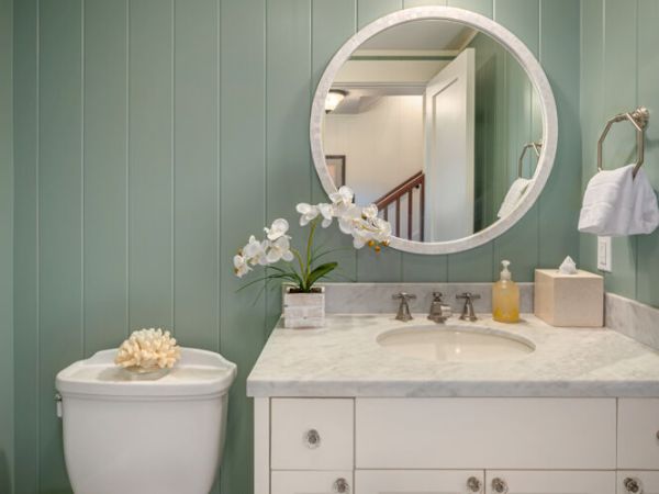 The image shows a bathroom with light green vertical panel walls, a round mirror, a sink with a white countertop, and a toilet with a decorative shell.