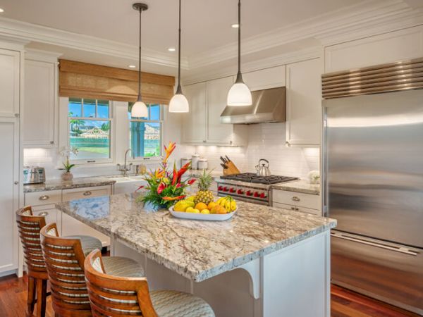 A modern kitchen featuring a large island with granite countertop, three stools, stainless steel appliances, and fresh flowers and fruit on the counter.