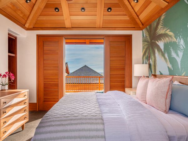 The image shows a tropical-themed bedroom with a wooden ceiling, a double bed, a dresser with flowers, and an open door leading to a balcony with a sea view.