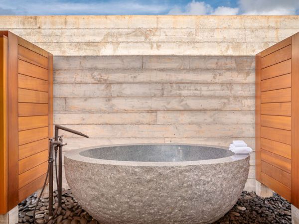 The image shows an outdoor stone bathtub with wooden privacy panels on either side, a faucet, and a stack of white towels against a concrete wall.