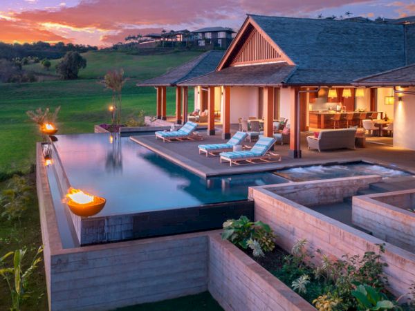 A luxurious villa with an infinity pool, lounge chairs, and fire features overlooking a scenic landscape, set against a beautiful sunset sky.