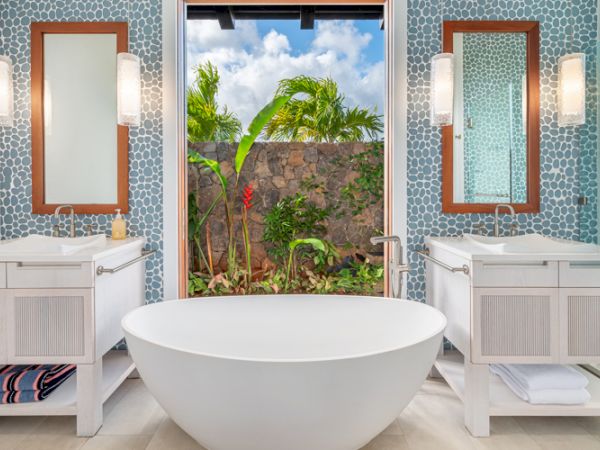 Modern bathroom with a standalone tub, twin sinks, mirrors, and windows revealing a lush, tropical garden view.