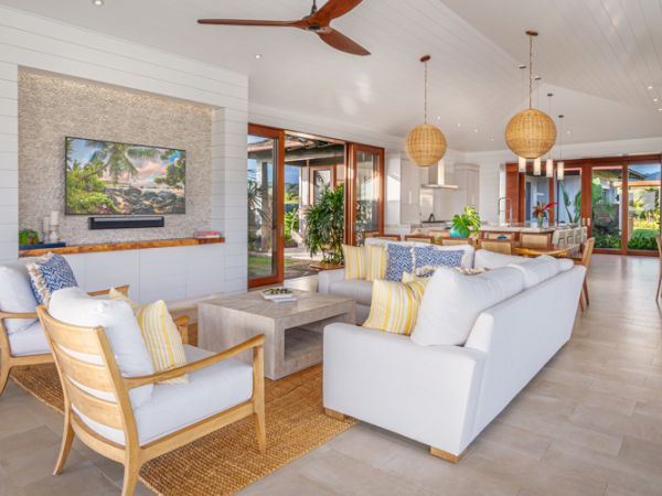 A spacious, modern living room with white sofas, wooden furniture, a mounted TV, ceiling fans, and large windows leading to an outdoor area.