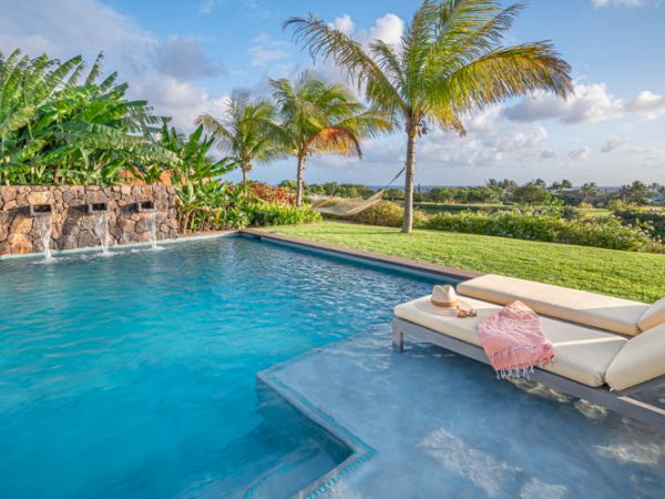 The image shows a serene pool with waterfalls, surrounded by lush greenery and palm trees, along with a comfortable lounge chair under a clear sky.