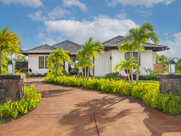 A driveway leading to a white single-story house surrounded by lush greenery and palm trees, with the address number 