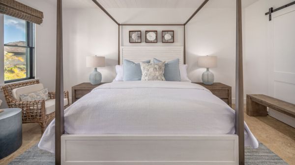 A stylish bedroom features a four-poster bed, bedside tables with lamps, a wicker chair, large window, and a sliding barn door.