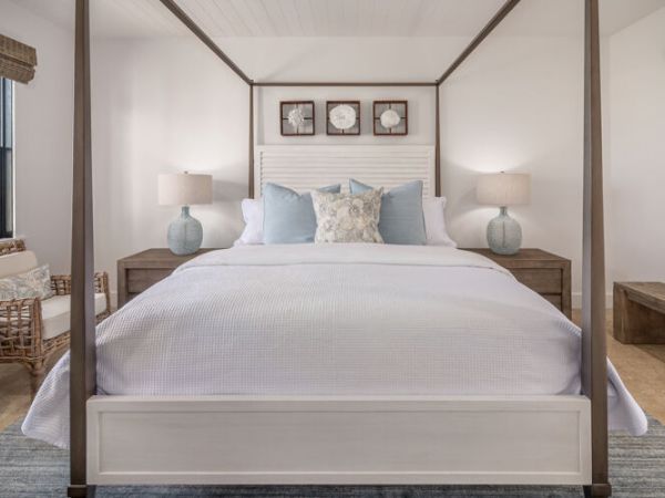 A stylish bedroom features a four-poster bed, bedside tables with lamps, a wicker chair, large window, and a sliding barn door.