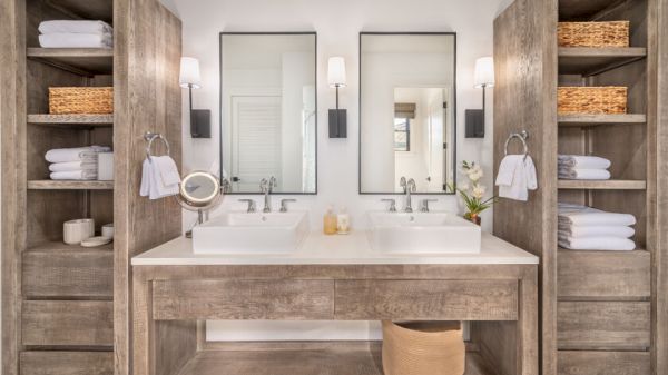A modern bathroom features dual sinks, wooden shelves with neatly folded towels, baskets, two mirrors, and wall-mounted lights, with a basket beneath them.