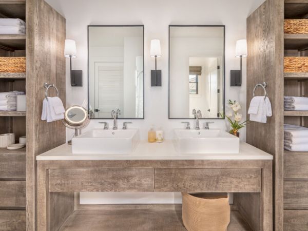 A modern bathroom features dual sinks, wooden shelves with neatly folded towels, baskets, two mirrors, and wall-mounted lights, with a basket beneath them.