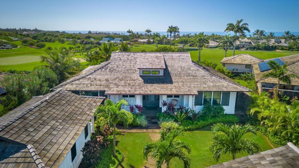 Aerial view of a bungalow-style house with a wooden roof, surrounded by lush greenery, palm trees, and neighboring houses, set against a backdrop of a golf course.