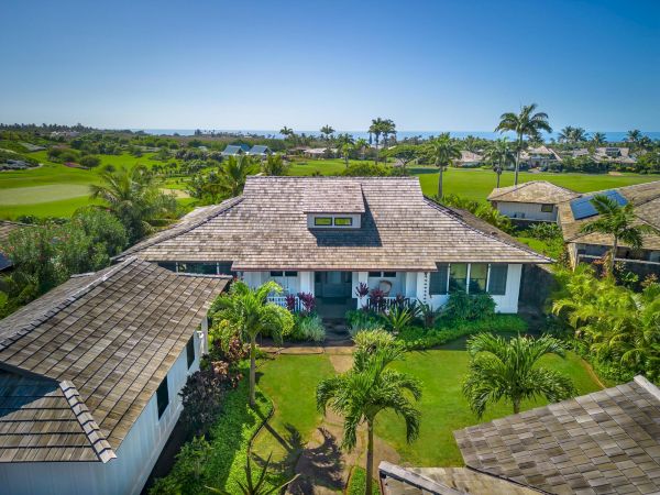 Aerial view of a bungalow-style house with a wooden roof, surrounded by lush greenery, palm trees, and neighboring houses, set against a backdrop of a golf course.