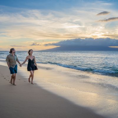A couple walking along a sandy beach at sunset, waves lapping the shore and a tranquil sky above.