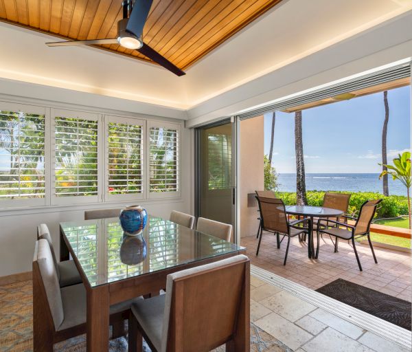 A bright dining area opens to a patio with ocean views, wooden ceiling, large windows, and a glass-top table with brown chairs.