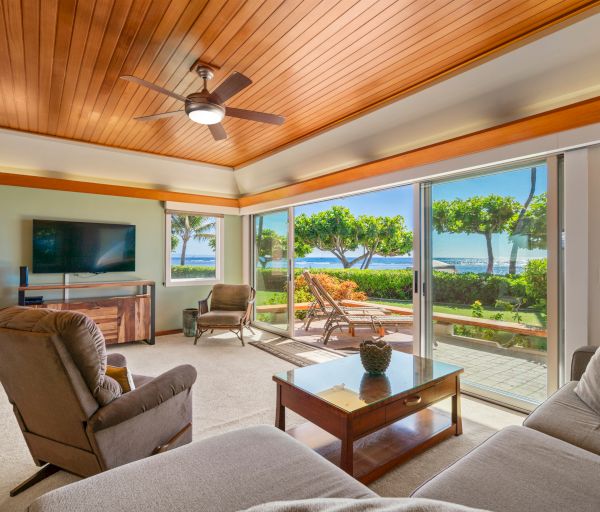 A bright coastal living room with tan couches, a wooden coffee table, ceiling fan, and a wall-mounted TV; large glass doors open to a sunny yard and sea view, creating a breezy, oceanfront vibe.