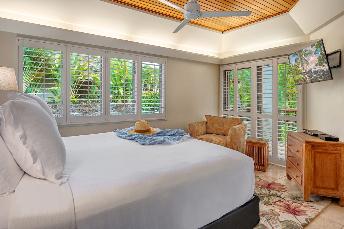 A cozy bedroom with a bed, hat, and scarf, adjacent to a window with blinds. There is a chair, TV, and tropical-themed d&eacute;cor. A door leads outside.