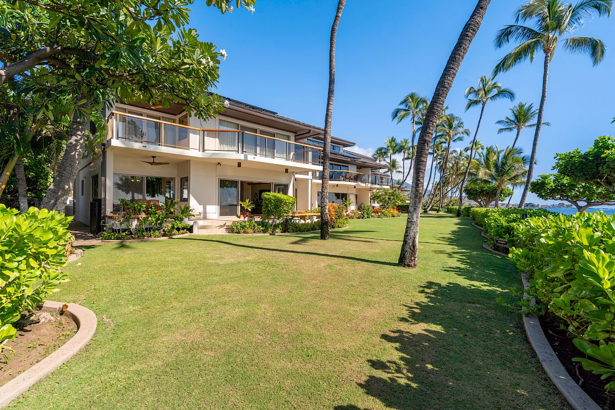 A tropical beachfront resort with a two-story building, palm trees, manicured lawn, hedges, and clear blue sky by the sea.