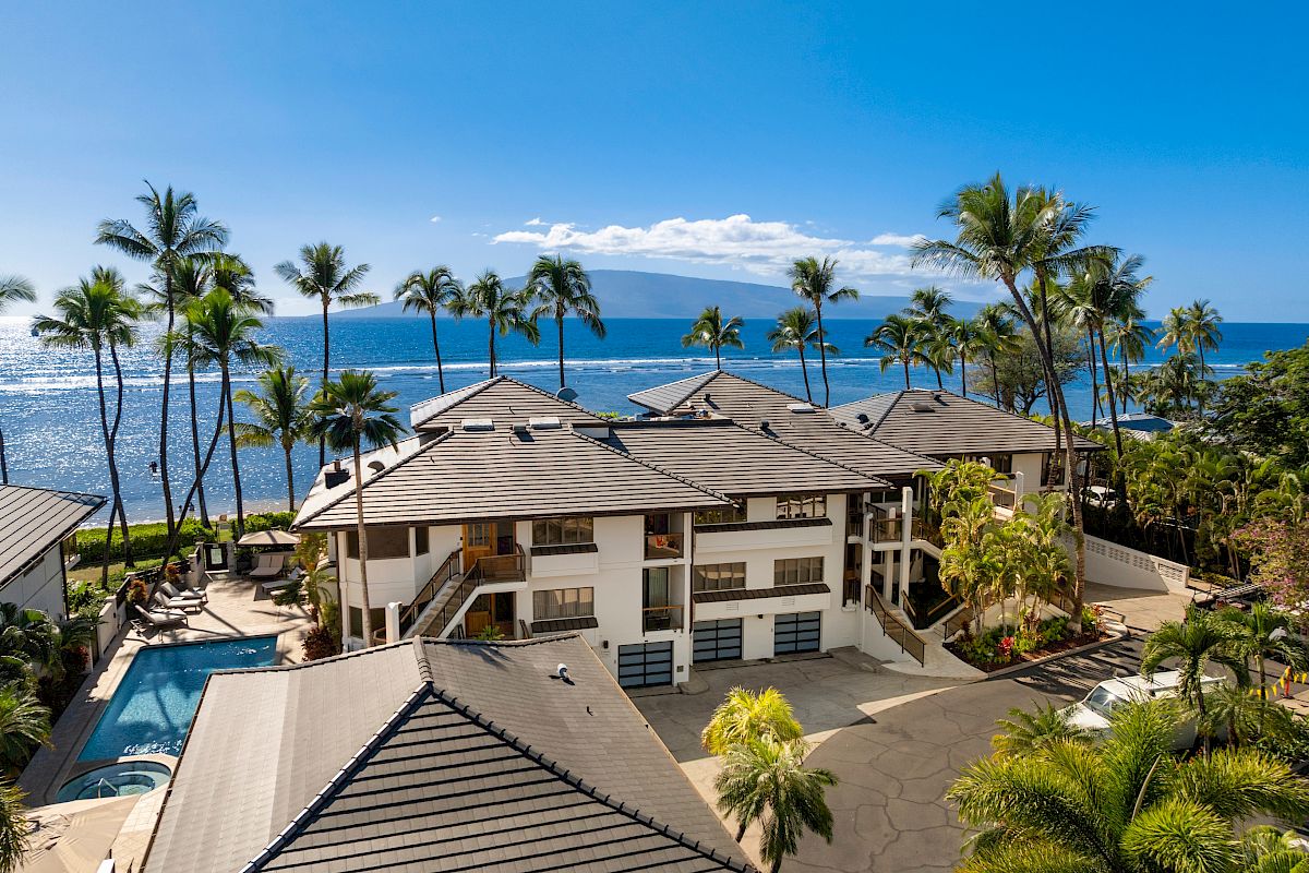 A tropical beachfront resort with modern villas, palm trees, a pool, and the ocean in the background under a bright blue sky.
